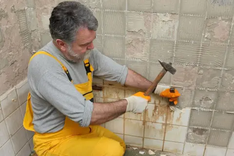 Un homme en salopette jaune utilise un marteau pour enlever du carrelage mural.