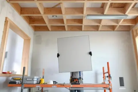 Un ouvrier sur un échafaudage s'apprête à poser une plaque pour faire un faux plafond. La structure en bois est visible.