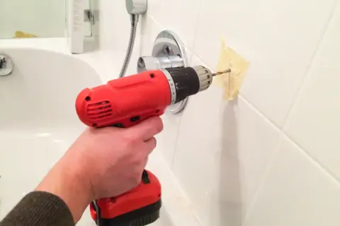 Une main utilise une perceuse pour percer du carrelage dans une salle de bain, avec du ruban adhésif pour guider le foret.
