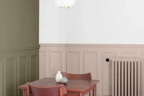 Peinture salon deux tons : mur vert olive contrastant avec un soubassement rose poudré orné de moulures. Une table en bois et des chaises sont disposées près d'un radiateur.