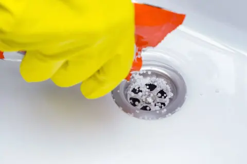 Gant jaune versant des granulés dans un siphon de douche pour déboucher la douche.