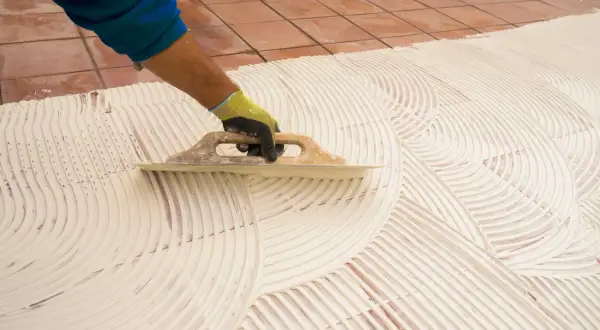 Main tenant, pose carrelage 60x60. Un artisan étale la colle avec une truelle pour préparer la pose de grands carreaux.