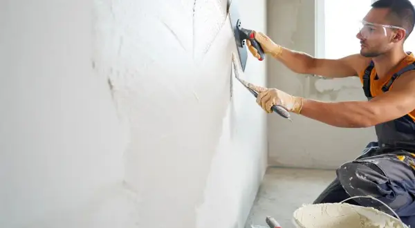 Un ouvrier en tenue de travail, lunettes de protection et gants, est en train de faire de l'enduit sur un mur avec une taloche et une truelle.