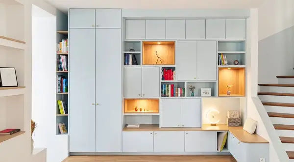 Un mur habillé de rangements modulables bleu pâle, avec des niches éclairées et une banquette intégrée près d'un escalier en bois.