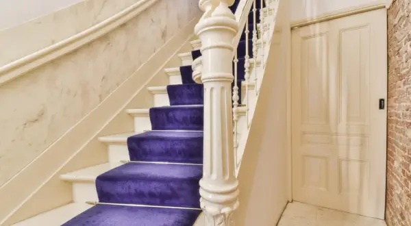 Escalier majestueux avec une moquette violette éclatante, un garde-corps blanc sculpté et un mur en marbre. Une porte crème et un mur de briques complètent la scène.