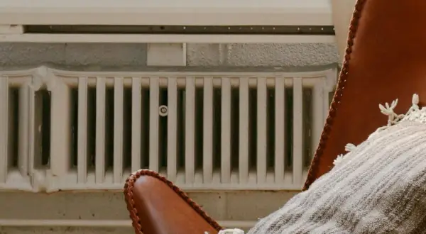 Un fauteuil en cuir marron avec un plaid douillet est placé près de vieux panneaux rayonnants blancs, créant une ambiance chaleureuse.