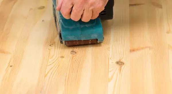 Main d'une personne utilisant une ponceuse pour poncer un parquet en bois clair.