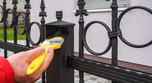 Maintenir le fer forgé propre avant peinture. Une main tient un pinceau jaune pour nettoyer la clôture.