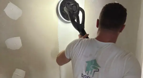 Un ouvrier utilise une ponceuse pour lisser un mur, préparant la surface pour poser bande placo.