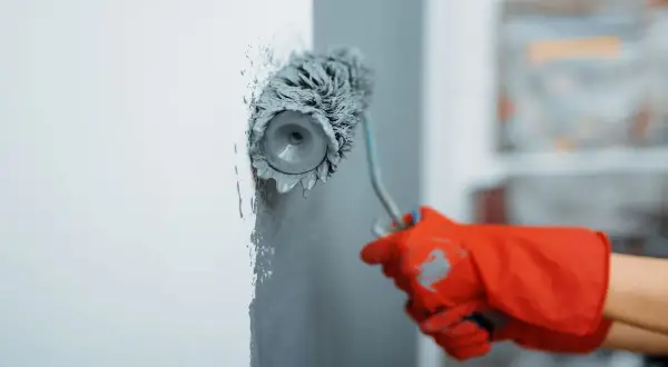 Maintenant, vous savez comment peindre un mur : une main gantée de rouge utilise un rouleau pour appliquer de la peinture grise sur un mur blanc.
