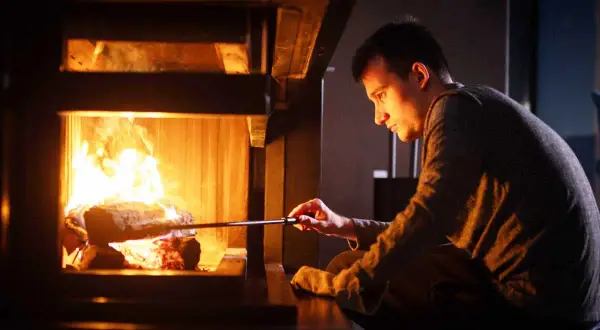 Un homme ajuste des bûches dans un poêle à bois, le feu crépite. Le tirage est parfait pour une soirée chaleureuse.