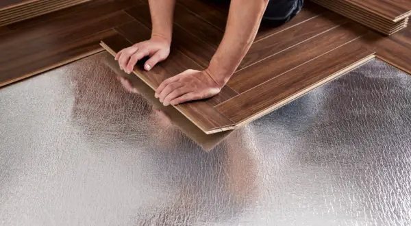 Pose bâton rompu : des mains ajustent une lame de parquet sur une sous-couche isolante argentée.