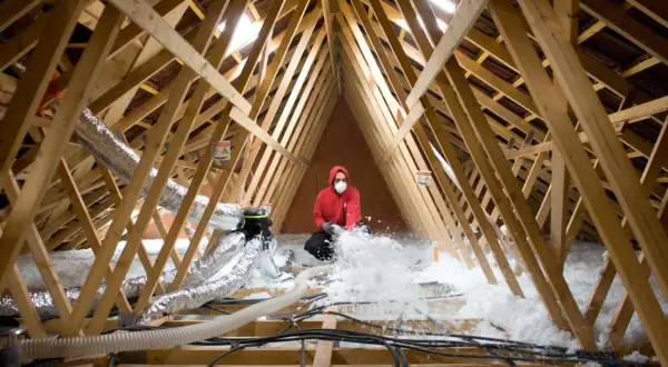 Un ouvrier en masque et sweat rouge réalise l'isolation des combles par soufflage, projetant de la ouate de cellulose dans la charpente.