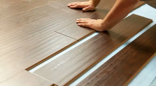 Pose de lames de parquet imitation bois sur une sous-couche bleue. L'isolation plancher bois est en cours, les mains guident l'assemblage.