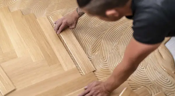 Pose d'un parquet flottant en chevrons. Un artisan applique la colle sur le sol avant de placer les lames de bois.