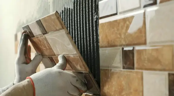 Mains gantées appliquant du mortier pour la pose carrelage d'une mosaïque murale aux tons marron et beige.