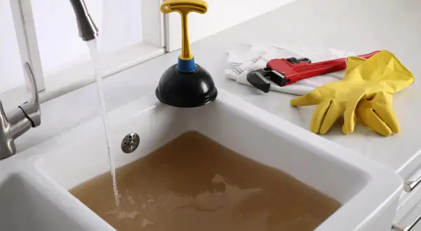 Évier bouché avec de l'eau sale. Un déboucheur, une clé à molette rouge et des gants jaunes sont prêts pour l'action.