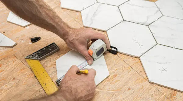 Mains mesurant une tuile hexagonale blanche sur un plancher OSB, avec des outils de pose.