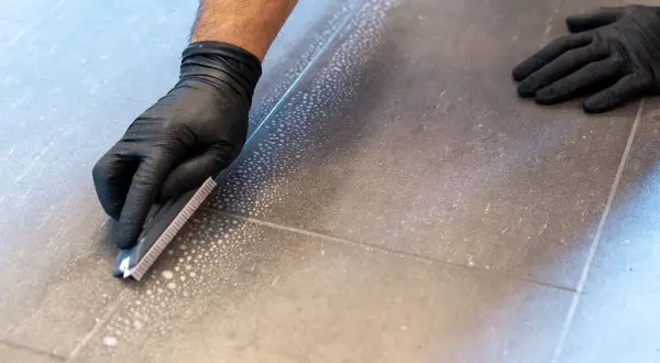 Main tenant enlever joint carrelage avec une spatule. Des bulles de savon indiquent le nettoyage.