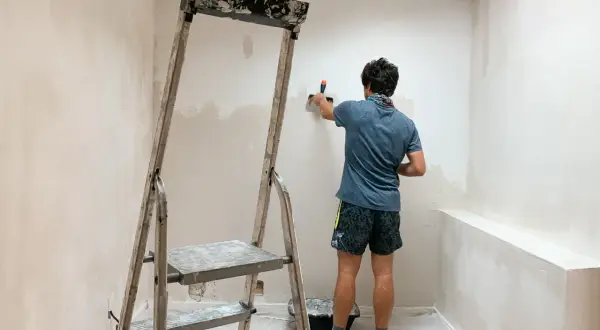 Un homme enlever du crépi intérieur sur un mur, à l'aide d'une spatule, près d'un escabeau.