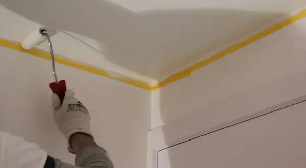 Main tenant en main un rouleau, il s'apprête à peindre un plafond. Le ruban de masquage jaune protège les bords.