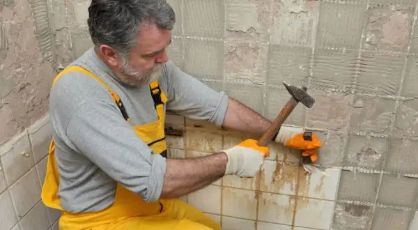 Un homme en salopette jaune utilise un marteau pour enlever du carrelage mural.