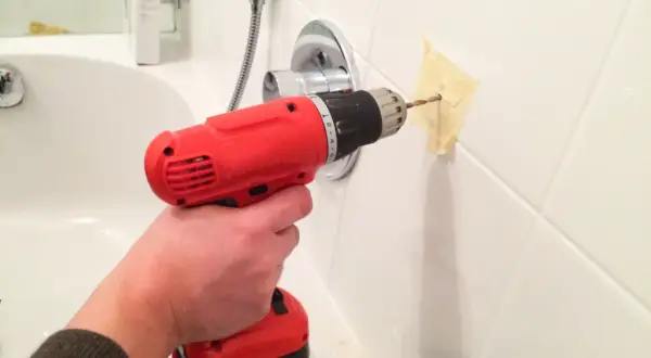 Une main utilise une perceuse pour percer du carrelage dans une salle de bain, avec du ruban adhésif pour guider le foret.