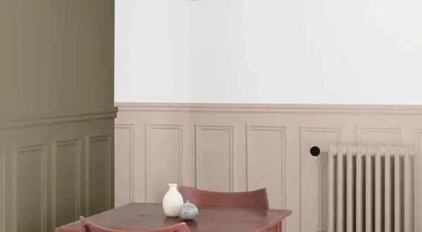 Peinture salon deux tons : mur vert olive contrastant avec un soubassement rose poudré orné de moulures. Une table en bois et des chaises sont disposées près d'un radiateur.