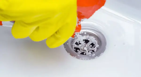 Gant jaune versant des granulés dans un siphon de douche pour déboucher la douche.