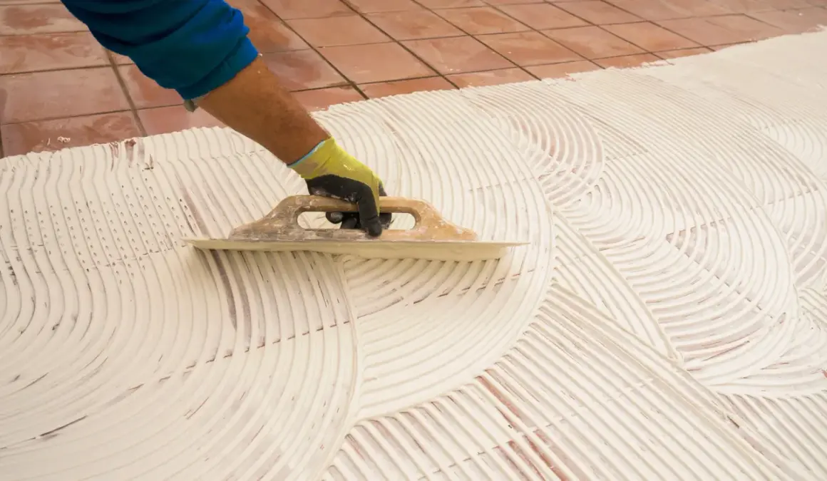 Main tenant, pose carrelage 60x60. Un artisan étale la colle avec une truelle pour préparer la pose de grands carreaux.