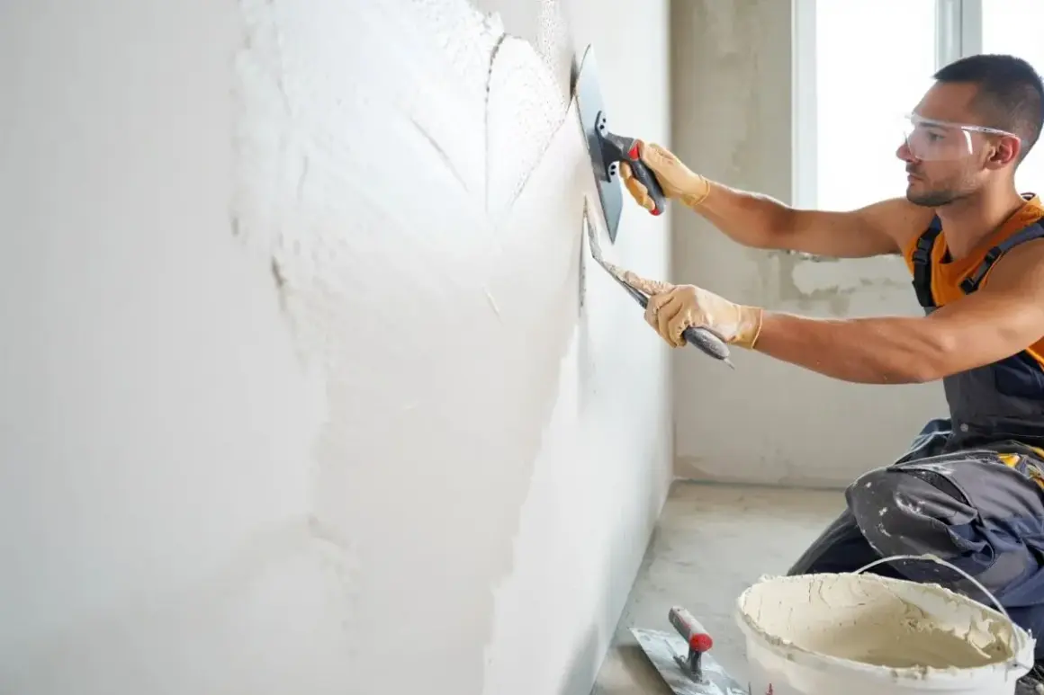 Un ouvrier en tenue de travail, lunettes de protection et gants, est en train de faire de l'enduit sur un mur avec une taloche et une truelle.