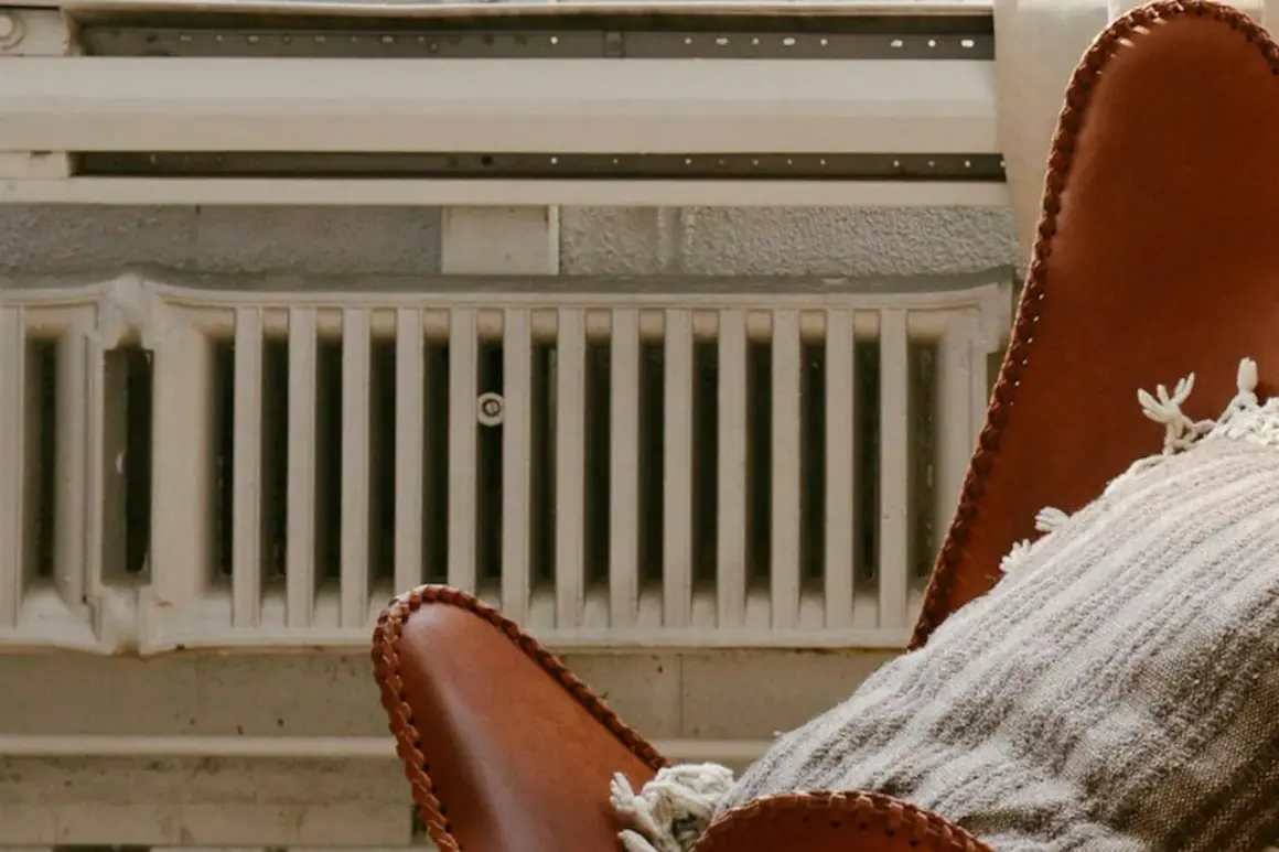 Un fauteuil en cuir marron avec un plaid douillet est placé près de vieux panneaux rayonnants blancs, créant une ambiance chaleureuse.