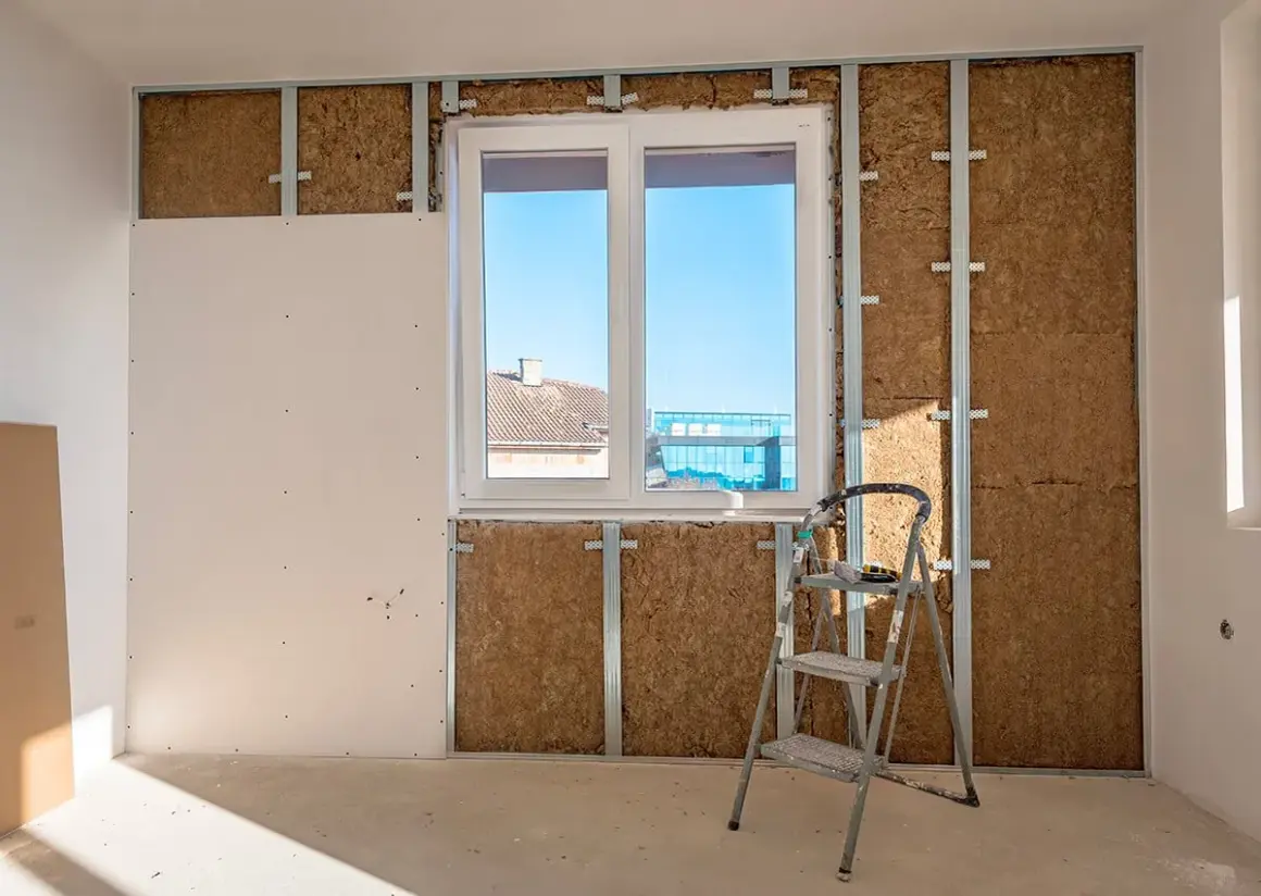 Travaux d'isolation intérieure : murs en cours de pose, avec laine de bois visible derrière une ossature métallique et une plaque de plâtre. Une échelle est présente.