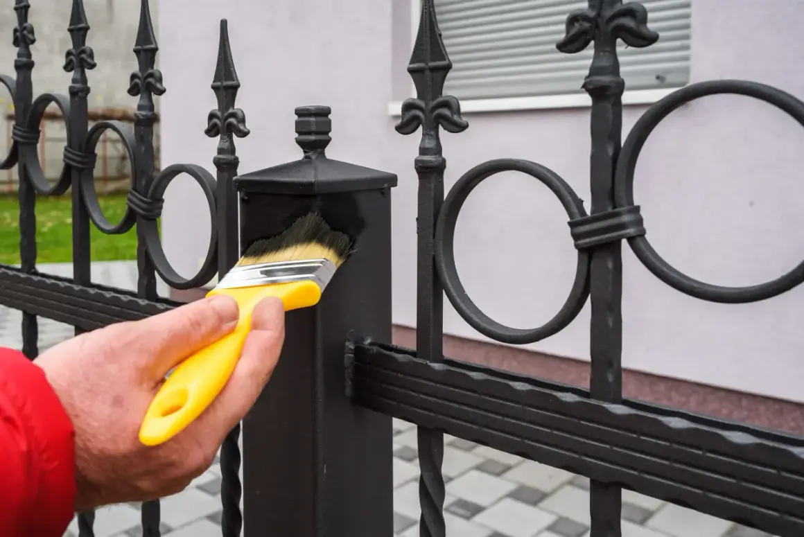Maintenir le fer forgé propre avant peinture. Une main tient un pinceau jaune pour nettoyer la clôture.