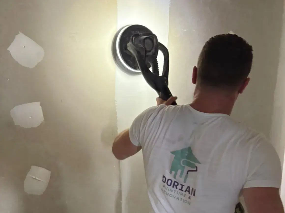 Un ouvrier utilise une ponceuse pour lisser un mur, préparant la surface pour poser bande placo.