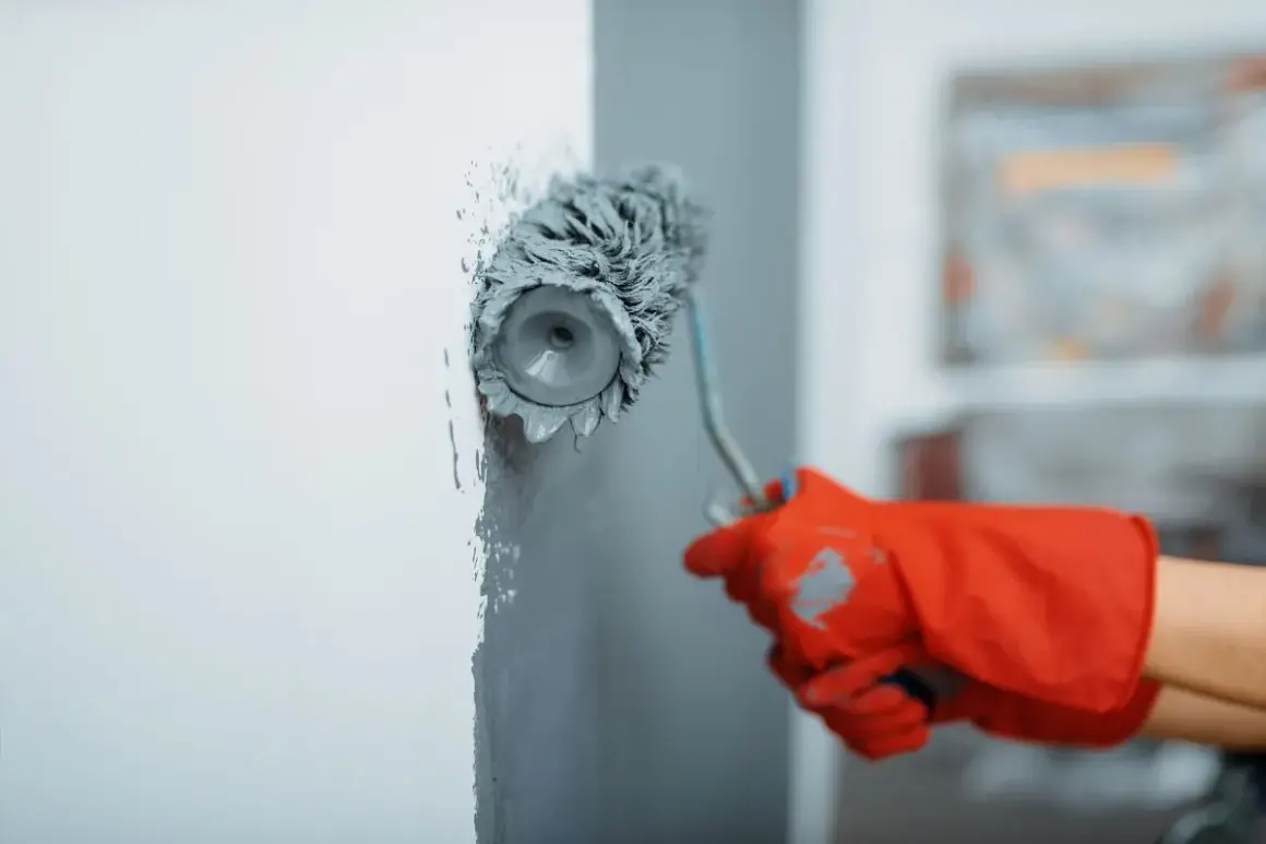 Maintenant, vous savez comment peindre un mur : une main gantée de rouge utilise un rouleau pour appliquer de la peinture grise sur un mur blanc.