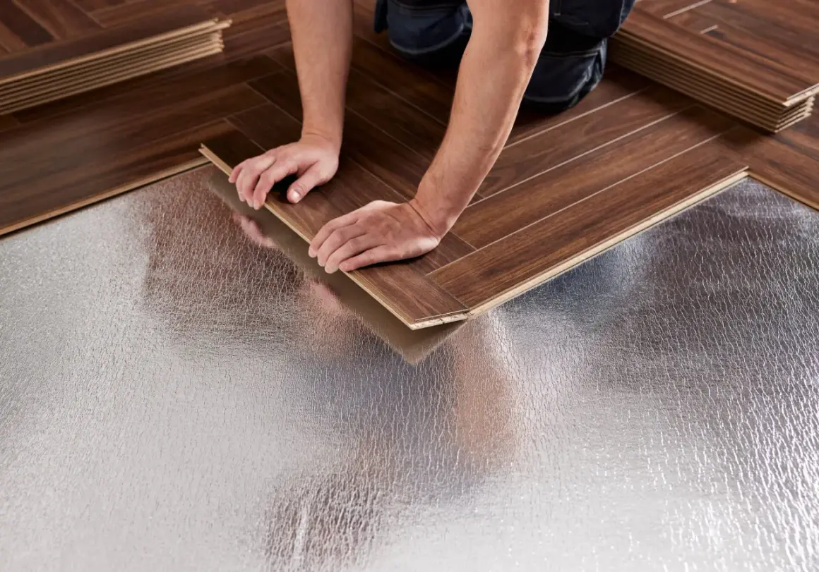 Pose bâton rompu : des mains ajustent une lame de parquet sur une sous-couche isolante argentée.