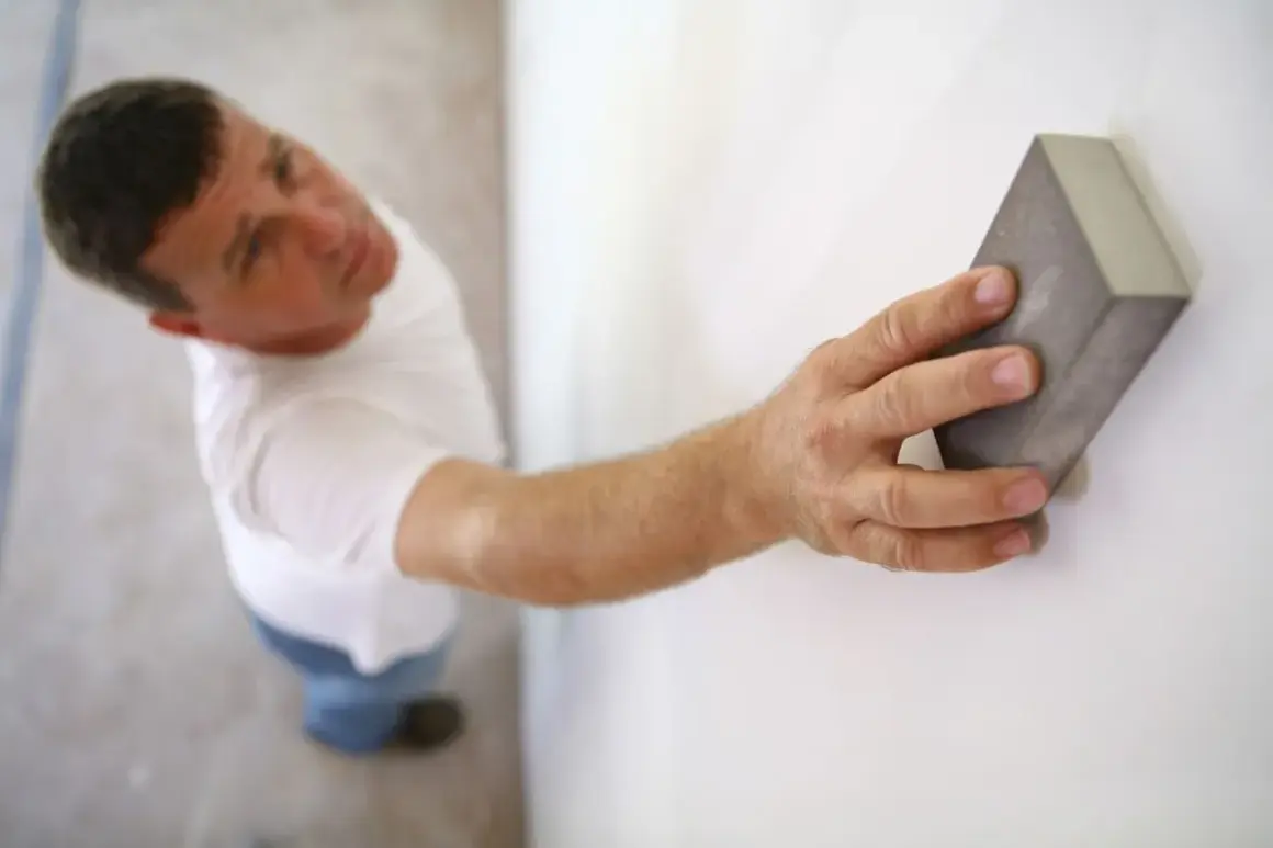 Un homme ponce un mur blanc avec une cale à poncer, préparant la surface avant de peindre.