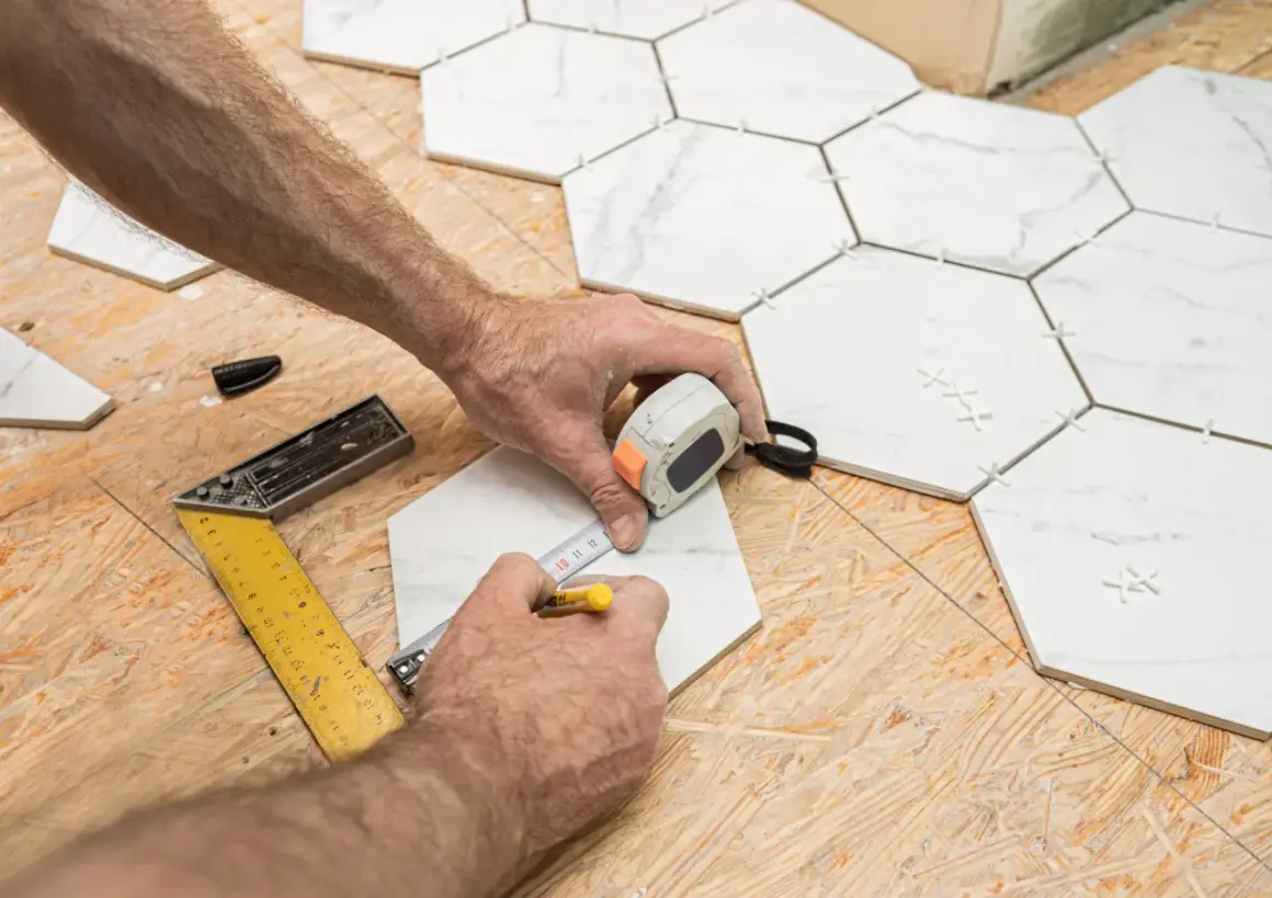 Mains mesurant une tuile hexagonale blanche sur un plancher OSB, avec des outils de pose.