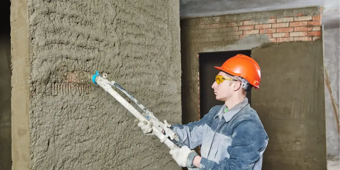 Un ouvrier en casque orange utilise un pistolet à crépir pour étanchéifier un mur en béton brut.