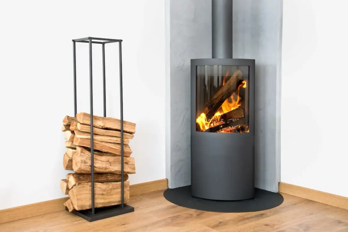 Un poêle à bois moderne avec un feu vif, à côté d'une pile de bois sur un support, le tout sur un mur en placo.