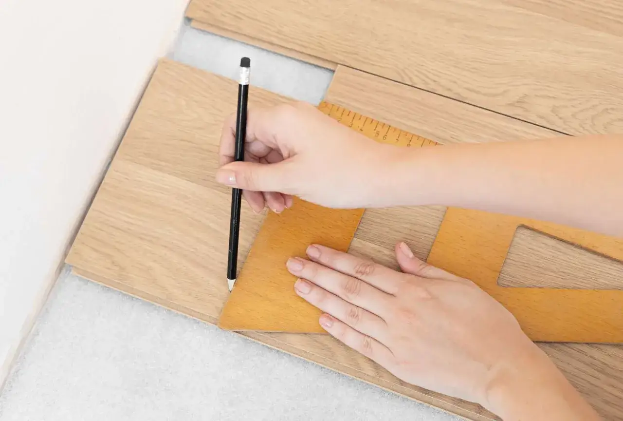Main d'une personne traçant une ligne sur une lame de parquet avec un crayon et une équerre pour une découpe précise.