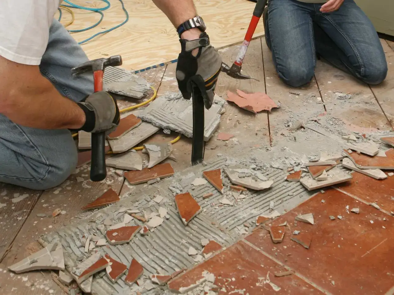 Deux personnes s'affairent à enlever du carrelage avec des marteaux et des burins, créant un chantier poussiéreux.