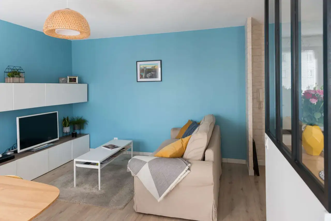 Un salon moderne aux murs bleu ciel, avec un canap&eacute; beige, une table basse blanche et une &eacute;tag&egrave;re murale. Id&eacute;es pour comment peindre un mur d&eacute;j&agrave; peint.