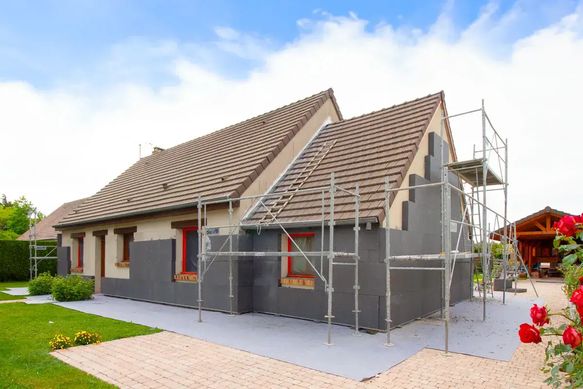 Maison en cours d'isolation extérieure avec panneaux gris, améliorant son inertie thermique. Échafaudages et roses rouges en premier plan.