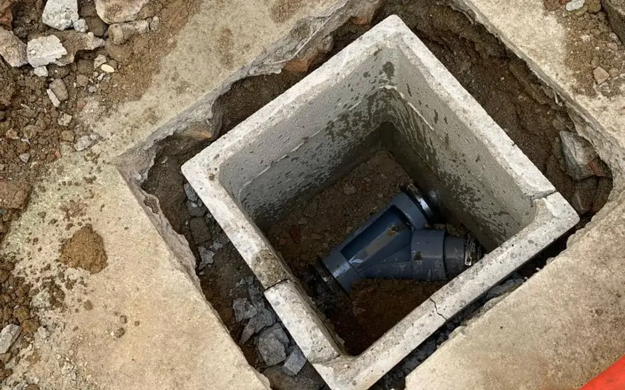 Un regard tout à l'égout en béton, avec des tuyaux gris à l'intérieur, entouré de terre et de gravats.