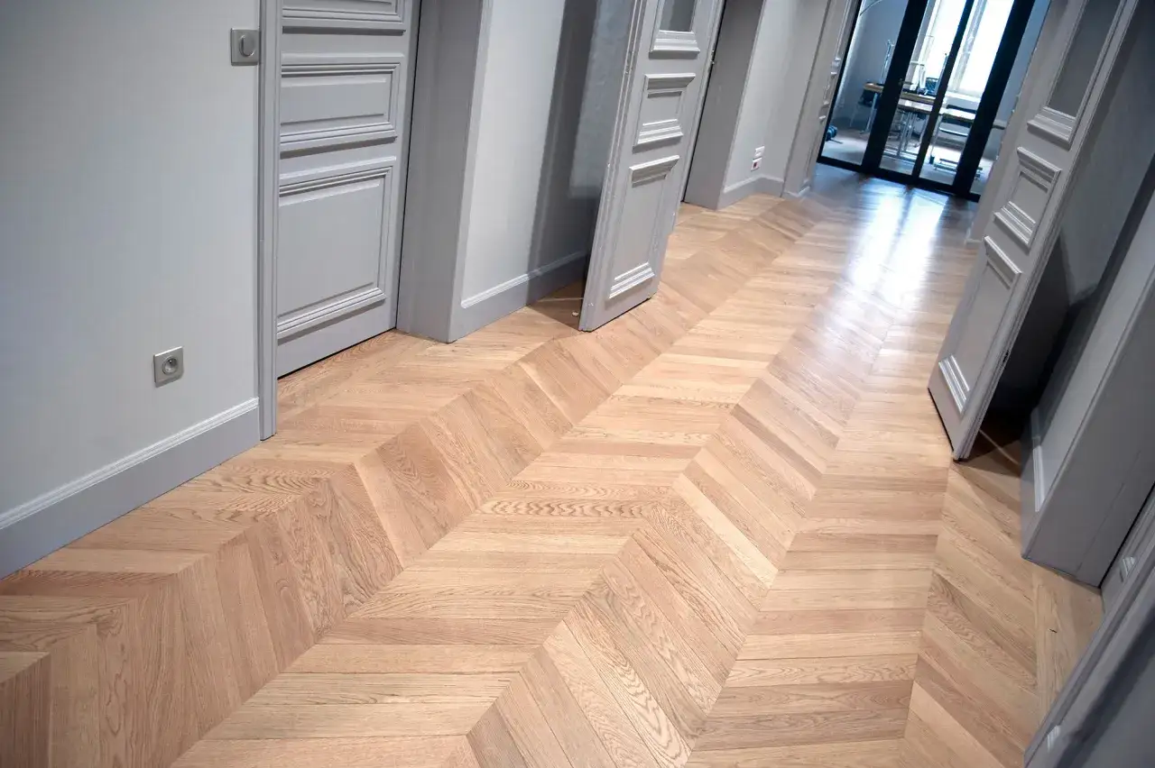 Un couloir élégant avec un pose parquet point de Hongrie en bois clair. Des portes grises aux moulures fines bordent le passage.