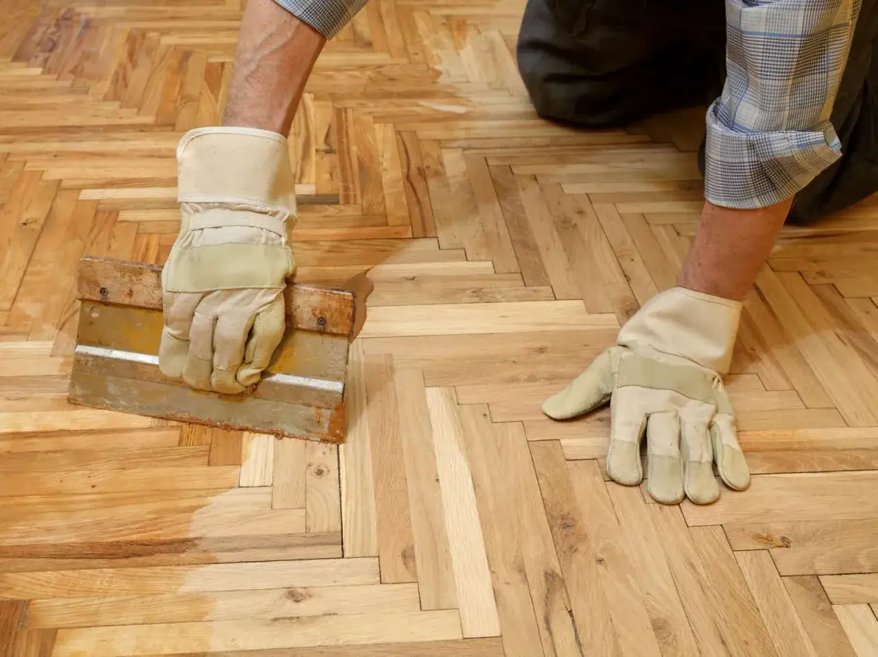 Application de la cire sur un parquet en chevrons avec une spatule. L'homme porte des gants pour comment cirer un parquet.