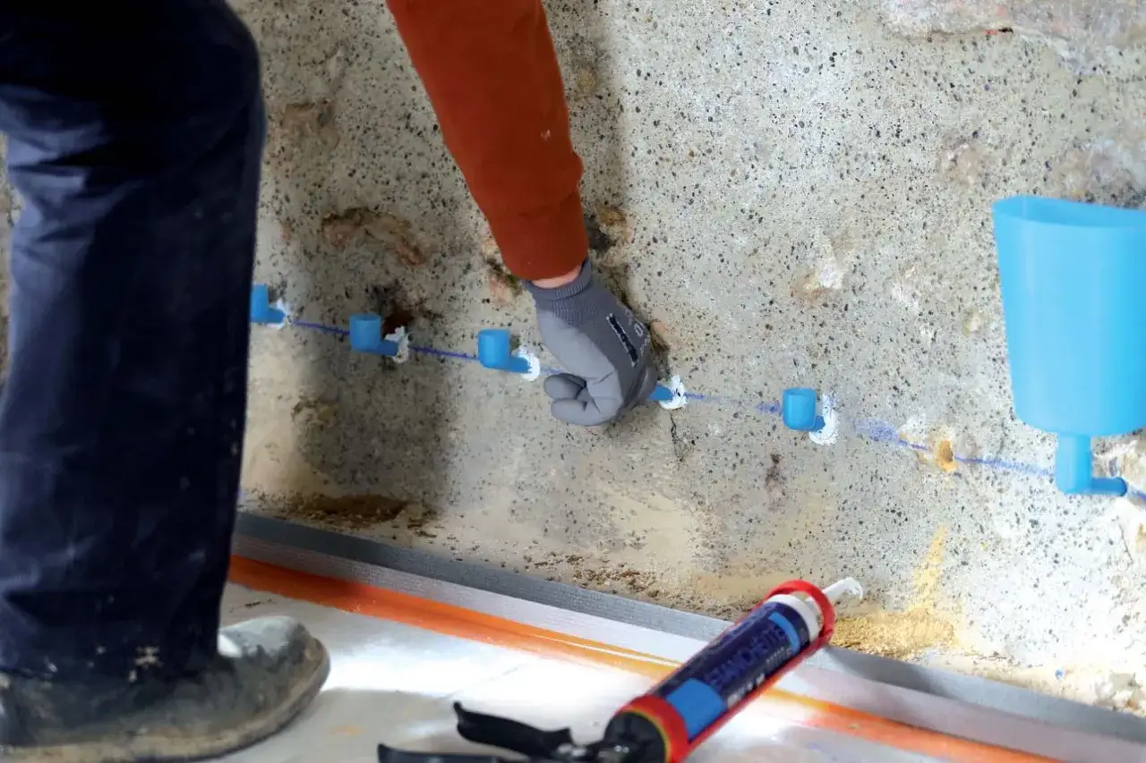 Un artisan utilise un pistolet &agrave; mastic pour &eacute;tanch&eacute;ifier un mur, injectant un produit dans des tubes bleus fix&eacute;s &agrave; la paroi.