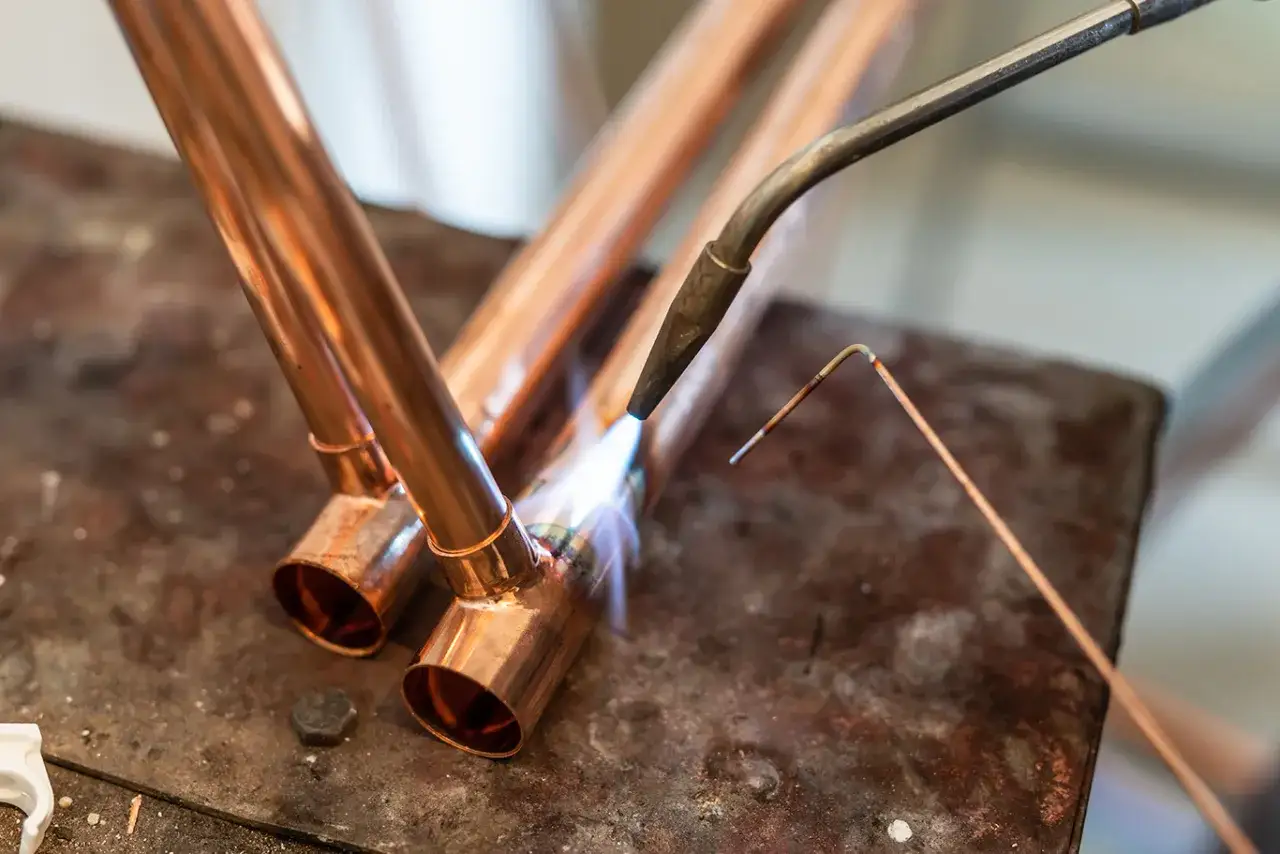 Un artisan r&eacute;alise une soudure cuivre plomberie sur des tuyaux de cuivre, la flamme du chalumeau illuminant la sc&egrave;ne.